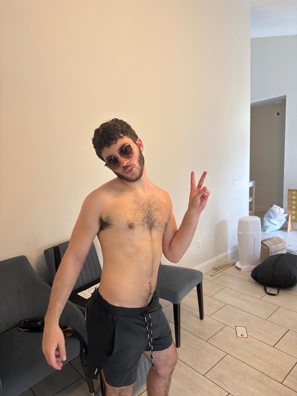 Sruly shirtless in sunglasses flashing a peace sign in a living room