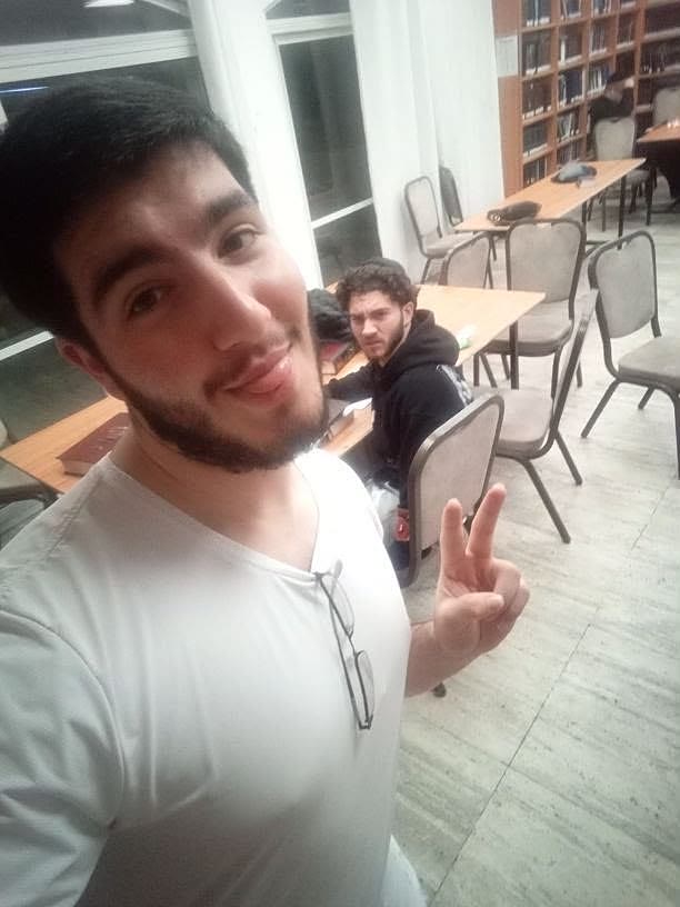 A friend taking a selfie in a library while Sruly glares at the camera from a table in the background