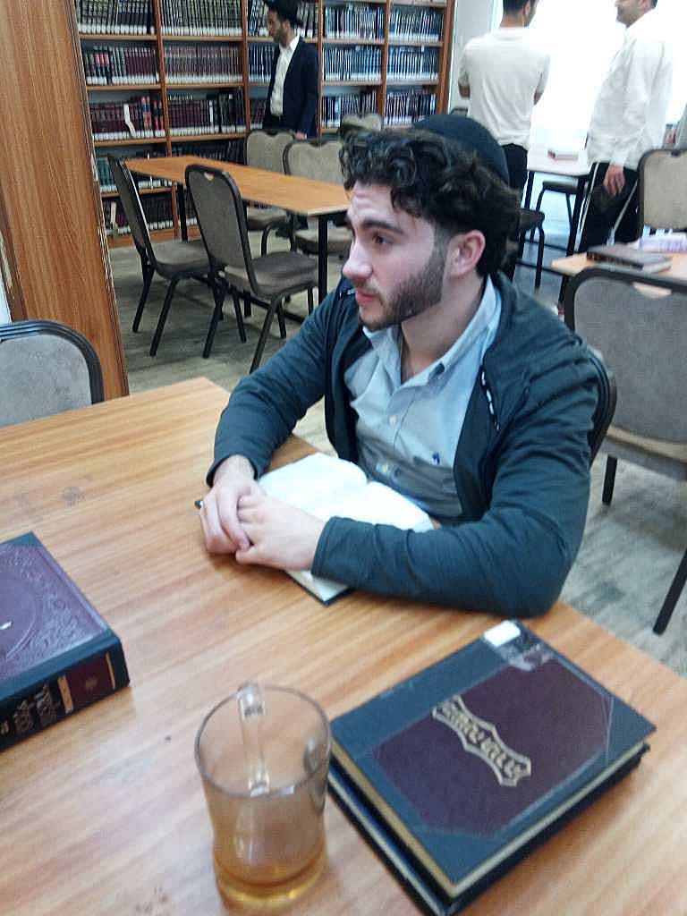 Sruly sitting at a library table with books, gazing thoughtfully into the distance