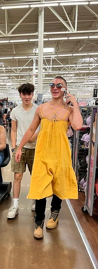 Sruly walking through Walmart in a yellow sundress, black pants, work boots and sunglasses, on the phone with a card taped to his face