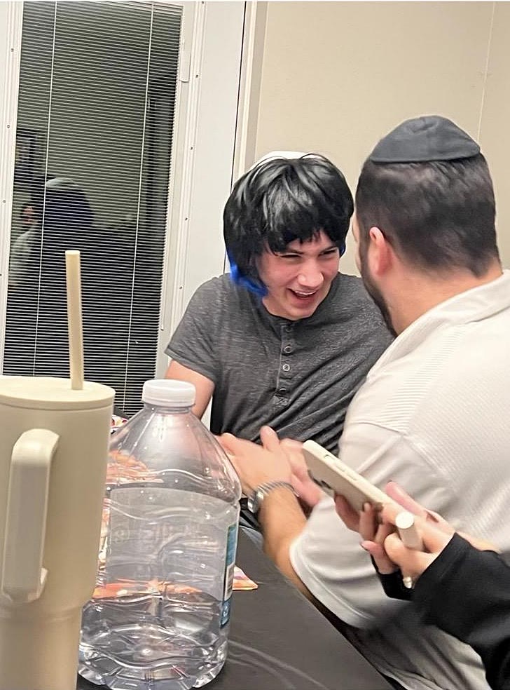 Sruly laughing in a black and blue wig while a friend shows him a phone